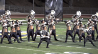 A marching band performs on at Cougar Stadium at College of the Canyons under stadium lights. The brass players, wearing matching ornate jackets and black pants, hold their instruments up in unison while two color guard members in contrasting silver and black outfits strike dynamic poses in front. The scene is highly synchronized and theatrical, with backdrops in the background and a dramatic sense of movement and energy.